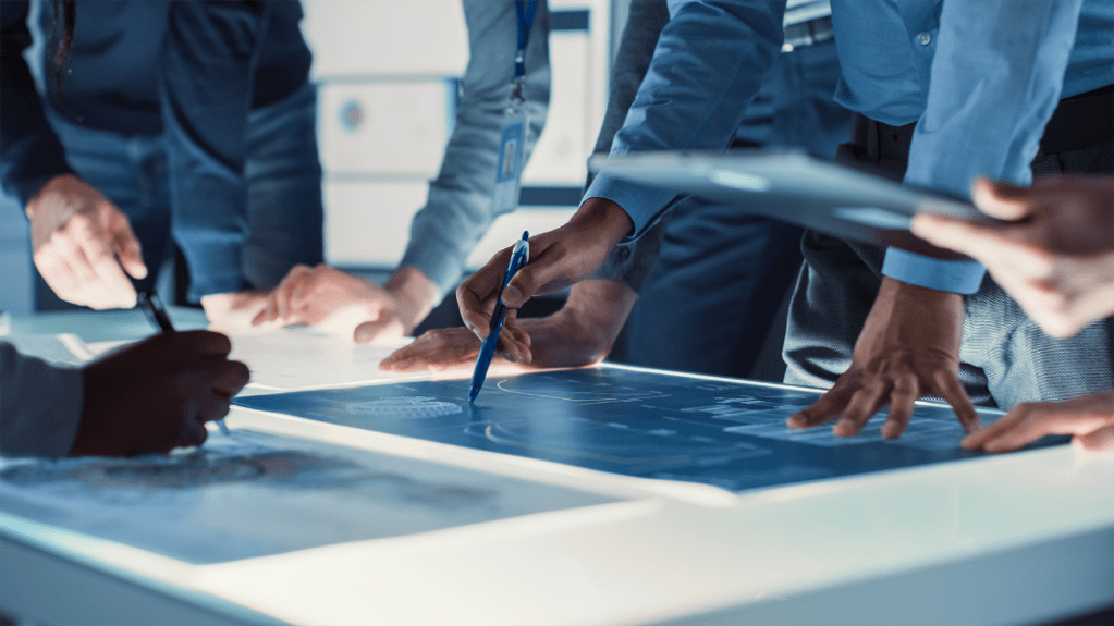 Multiple people standing over a blueprint with the main subject holding a pencil
